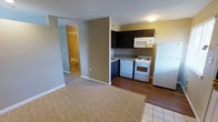 This single-floor unit offers a practical and well-organized layout designed for comfortable everyday living. The open-concept design connects the living area and kitchen seamlessly, creating a cohesive and functional space. The kitchen is equipped with white appliances, including a refrigerator, electric range with oven, and an over-the-range microwave. Dark espresso-toned cabinetry provides generous storage and contrasts nicely with the granite-look countertops. The kitchen features durable hardwood-style flooring, while the living area transitions to a warm carpet, adding comfort underfoot. Neutral yellow and grey tones on the walls contribute to a bright and welcoming ambiance throughout the unit. A hallway connects the main living space to a bathroom, which is visible in the background and features light-toned finishes. The layout makes efficient use of the available space, with clean lines and white baseboards adding a crisp finishing detail to each room. Overall, the unit presents a tidy, move-in-ready interior with a cohesive color palette and practical finishes suited for a range of lifestyles.