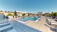 This rooftop amenity deck offers a well-appointed outdoor retreat designed for relaxation and socializing. At its centerpiece is a resort-style swimming pool with a distinctive geometric shape, featuring tiled finishes in cool blue tones and accessible entry equipment. Surrounding the pool, a generous sun deck is lined with sleek lounge chairs, providing ample space for sunbathing under open skies. A bold color palette of vibrant orange Adirondack chairs and coordinating furniture adds energy and personality to the space. A prominent wood pergola anchors the social seating area, its warm timber beams providing partial shade while framing views of the surrounding cityscape. Beneath the pergola, casual seating arrangements invite conversation and relaxation, complemented by colorful accent tables and chairs. Large sliding glass doors connect the outdoor deck to an interior space, allowing for seamless indoor-outdoor flow. The entire deck is finished with large-format light gray tiles, lending a clean, contemporary aesthetic throughout. Lush planted greenery in raised beds softens the urban edges, while an outdoor screen adds an entertainment element. The open-air setting enjoys abundant natural light and sweeping rooftop views, making this a dynamic and inviting communal space.