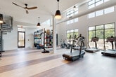 This well-appointed fitness center is designed to support a full range of workout routines in a bright, open environment. The space is defined by soaring double-height ceilings lined with a row of clerestory windows that flood the interior with natural light throughout the day. Large floor-to-ceiling windows along one wall offer views of the outdoor surroundings, including what appears to be a pool area, further enhancing the airy atmosphere. Warm-toned hardwood flooring runs throughout, adding a polished, modern aesthetic that contrasts with the industrial-style exposed ductwork above.

The cardio zone is well-equipped with multiple Cascade treadmills, elliptical machines, and stationary bikes, all positioned to take advantage of the outdoor views. A dedicated strength training area features a multi-functional cable machine, a squat rack with bench, and a full rack of dumbbells. A functional training wall organizes an extensive collection of medicine balls, resistance equipment, and accessories for dynamic workouts.

Pendant lighting and ceiling fans complement the natural light, maintaining comfort during intense sessions. A striking large-scale mural adds an artistic focal point to one section of the space. A sculptural spiral staircase in matte black serves as both a functional and architectural element. The overall ambiance is clean, modern, and energizing.