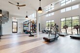 This well-appointed fitness center is designed to support a full range of workout routines in a bright, open environment. The space is defined by soaring double-height ceilings lined with a row of clerestory windows that flood the interior with natural light throughout the day. Large floor-to-ceiling windows along one wall offer views of the outdoor surroundings, including what appears to be a pool area, further enhancing the airy atmosphere. Warm-toned hardwood flooring runs throughout, adding a polished, modern aesthetic that contrasts with the industrial-style exposed ductwork above.

The cardio zone is well-equipped with multiple Cascade treadmills, elliptical machines, and stationary bikes, all positioned to take advantage of the outdoor views. A dedicated strength training area features a multi-functional cable machine, a squat rack with bench, and a full rack of dumbbells. A functional training wall organizes an extensive collection of medicine balls, resistance equipment, and accessories for dynamic workouts.

Pendant lighting and ceiling fans complement the natural light, maintaining comfort during intense sessions. A striking large-scale mural adds an artistic focal point to one section of the space. A sculptural spiral staircase in matte black serves as both a functional and architectural element. The overall ambiance is clean, modern, and energizing.