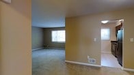 This single-story space presents an open, functional layout characterized by warm, neutral-toned walls that create an inviting atmosphere throughout. The living areas are finished with wall-to-wall carpeting in a neutral hue, providing a comfortable and cohesive feel underfoot. The space is divided by a partial wall, subtly defining separate zones while maintaining an open, connected flow between areas.

Natural light enters through a window fitted with white horizontal blinds, offering a view of surrounding greenery outside. White baseboards line the perimeter of the rooms, providing a clean contrast against the warm tan walls. Floor-level HVAC vents are positioned throughout, ensuring efficient climate control across the space.

A light fixture is visible in an adjoining area, suggesting additional functional zones beyond the main living space. The layout appears practical and straightforward, with wall switch plates positioned at standard heights. Overall, the space conveys a clean, move-in-ready environment with a simple, understated aesthetic well-suited to a variety of furnishing styles.