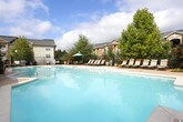 This single-level outdoor amenity space serves as a communal gathering area within a residential community. The patio is enclosed by a sturdy black iron fence with a gated entrance, providing a defined and secure boundary while maintaining open views of the surrounding landscaped grounds and multi-story residential buildings beyond.

The seating area features a round glass-top table paired with slatted chairs, shaded by a vibrant teal patio umbrella — ideal for outdoor dining or relaxed conversation. The space is accented with large decorative ceramic planters filled with bright red flowering plants, adding color and a welcoming aesthetic throughout.

A dedicated gas grill station, clearly marked with posted usage guidelines, is accessible from the patio and provides a convenient outdoor cooking amenity for residents. Brick detailing at the base of the building exterior complements the neutral siding and traditional-style doors, contributing to the classic architectural character of the space.

Wall-mounted lantern-style light fixtures are positioned along the building facade, ensuring the area remains functional into the evening hours. The concrete patio surface is clean and well-maintained, and additional seating is available along the building's covered section, offering a shaded retreat. Overall, this outdoor space provides a comfortable and practical environment for community use.