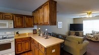 This two-floor home showcases a thoughtfully designed open-concept layout that seamlessly connects the kitchen, dining, and living areas. The kitchen is a standout feature, outfitted with rich, warm-toned wood cabinetry that extends to the ceiling, providing generous storage throughout. A peninsula with a light-colored countertop serves as both a functional workspace and an informal breakfast bar, complete with built-in outlets for added convenience. Decorative candle accents on the peninsula contribute to a cozy atmosphere.

Adjacent to the kitchen, the dining area accommodates a full table and chair set, flowing naturally into the living room beyond. The living room is comfortably furnished, featuring a media console with a flat-screen television and a large sofa, creating a relaxed and inviting space. Neutral gray walls throughout the open-concept area complement the warm wood tones of the cabinetry and hardwood-style flooring visible in the kitchen zone. A textured ceiling adds a layer of character to the space. The overall ambiance is warm, casual, and functional, making it well-suited for everyday living and entertaining.