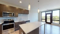 This contemporary single-floor residence combines modern design with functional living. The open-concept kitchen serves as the heart of the space, showcasing rich dark-toned cabinetry with brushed gold hardware, a white quartz countertop island with pendant lighting overhead, and a full suite of stainless steel appliances including a refrigerator, range, microwave, and dishwasher. A subway tile backsplash adds a clean, classic finish, while light hardwood flooring runs throughout the main living areas, lending warmth and continuity. The kitchen opens toward a sliding glass door that leads to a private balcony, offering views of surrounding greenery and filling the space with natural light. The bedroom is carpeted for comfort, featuring neutral tones and a window fitted with blinds for privacy and light control. The bathroom is finished with a dark wood vanity, white quartz countertop, a large mirror, and brushed nickel sconce lighting, maintaining the cohesive modern aesthetic found throughout the residence. A dedicated laundry room houses a full-size washer and dryer with overhead wire shelving for additional storage. The overall design reflects a clean, contemporary sensibility with quality finishes, practical storage, and thoughtful details throughout.