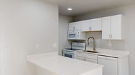 This thoughtfully updated upper-floor unit offers a clean, modern aesthetic throughout its efficiently designed layout. The kitchen serves as a central highlight, featuring white shaker-style cabinetry with brushed gold hardware, crisp white quartz countertops, a peninsula with additional prep and storage space, and a sleek industrial-style faucet over a stainless steel sink. White appliances, including an electric range, over-the-range microwave, and dishwasher, complete the space. Recessed lighting brightens the interior throughout. The living area is anchored by warm-toned hardwood-style flooring and leads to a private balcony through a sliding glass door, offering serene views of a landscaped courtyard framed by mature deciduous trees. Natural light filters generously into the living space, enhancing the airy feel. The bathroom is finished with a frameless glass walk-in shower, white subway tile surround, a single-sink vanity with white cabinetry and quartz countertop, and modern light fixtures above a framed mirror. Neutral gray walls and consistent finishes tie the space together, creating a cohesive and functional living environment with thoughtful attention to detail.