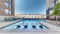 Located at 1900 S Acoma St in Denver's Baker neighborhood, this outdoor pool amenity offers a well-appointed resort-style retreat set within a modern urban courtyard. The centerpiece is a generously sized lap pool with crystal-clear blue water, surrounded by large-format tile decking and contemporary white chaise lounges perfect for sunbathing. Sleek in-pool loungers allow residents to relax partially submerged while soaking in the open Colorado sky.

Adjacent to the main pool is a spa featuring a striking stone water wall accent, with dual cascading jets framed by natural stacked stone masonry that contrasts beautifully with the surrounding contemporary architecture. Lush landscaped garden beds with ornamental trees, evergreen shrubs, and decorative rock accents are thoughtfully integrated throughout the courtyard, softening the urban setting.

A dedicated patio lounge area provides shaded outdoor dining beneath bold striped umbrellas, with colorful seating arrangements ideal for socializing. The space is enclosed by the building's modern façade, which incorporates brick, panel, and glass elements rising several stories above. Accessible entry points and railings ensure ease of use for all. The overall ambiance is vibrant yet relaxed, offering a true outdoor escape in the heart of Denver.
