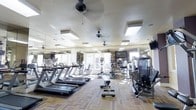 This fitness center offers a comprehensive workout environment designed to accommodate a wide range of exercise needs. The space is laid out on a single floor and features an open, airy feel enhanced by recessed skylights and large windows that invite abundant natural light. Ceiling fans are evenly distributed throughout, ensuring comfortable airflow during workouts.

The cardio zone is prominently featured along one side of the room, lined with multiple treadmills and upright stationary bikes, all facing toward the windows to provide an engaging view during exercise. Toward the center and rear of the space, additional cardio equipment such as ellipticals and cycle machines are thoughtfully arranged for easy access.

The strength training area is equipped with a selection of resistance machines, catering to users focused on muscle conditioning and toning. Free-standing benches and step platforms are also available for versatile functional training.

Floor-to-ceiling mirrors line the walls, visually expanding the space and allowing users to monitor their form throughout their workouts. Mounted flat-screen televisions provide entertainment and motivation. The flooring features a durable, patterned carpet tile that adds both comfort underfoot and a clean, polished aesthetic to the overall environment.