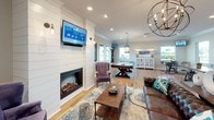 This single-story space is designed with an open-concept floor plan that seamlessly connects its key living areas, all unified by warm hardwood flooring and crisp crown molding throughout. The kitchen is a standout feature, showcasing gray shaker-style cabinetry, stainless steel appliances, and a striking arabesque tile backsplash. A large central island with a white quartz countertop provides ample workspace and bar seating, illuminated by a trio of pendant lights in amber glass. Natural light filters in through oversized windows, offering views of the surrounding landscape.

Adjacent to the kitchen, a cozy dining nook is framed by two windows and features a wall-mounted television, making it an ideal casual gathering spot. The living area is anchored by a shiplap-clad fireplace wall with built-in sconce lighting and a mounted television, creating a warm, farmhouse-inspired focal point. Comfortable seating and an ornate chandelier add character to the space.

The layout extends into a spacious game room complete with a billiard table, additional seating, and display shelving — perfect for entertainment. Throughout the space, neutral tones, recessed lighting, and consistent design elements create a cohesive, inviting ambiance.