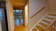 This two-storey space is defined by its warm, inviting character and classic design elements. The main level showcases distinctive parquet hardwood flooring laid in a geometric pattern, adding texture and visual interest underfoot. Natural light pours in generously through large floor-to-ceiling sliding glass doors fitted with vertical blinds, opening the living area to verdant outdoor views and creating a seamless connection between interior and exterior. The neutral-toned walls throughout contribute to a bright, open atmosphere that complements the natural wood tones of the flooring.

A hallway on the main level provides a sense of separation between the living areas, with a closet conveniently accessible nearby. The wood-railed staircase with matching wooden treads leads to the upper floor, maintaining a consistent warmth and continuity in materials throughout the home. The overall aesthetic is practical yet comfortable, with the combination of natural wood finishes and ample natural light creating an inviting ambiance suited to everyday living across both levels.