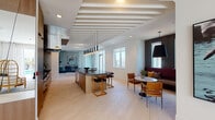 This elegantly designed single-floor space combines modern aesthetics with thoughtful functionality across a generous, open layout. Light-toned large-format tile flooring flows seamlessly throughout, unifying the distinct zones of the space.

The kitchen is outfitted with stainless steel appliances, dark stone countertops, a white subway tile backsplash, and warm wood cabinetry. A central island with gold-toned metal legs and bar seating serves as both a culinary workspace and a social focal point, anchored above by a dramatic linear pendant light arrangement suspended from a striking slatted ceiling feature.

Adjacent to the kitchen, a cozy dining nook is defined by built-in burgundy upholstered banquette seating, dark wood-paneled walls, a marble-top round table, and a bold oversized pendant light — framing city skyline views through flanking windows.

The expansive living area is bathed in natural light through floor-to-ceiling windows and French doors that open to an outdoor terrace overlooking a pool and the surrounding cityscape. Soaring slatted ceilings and a sculptural multi-arm chandelier add architectural drama to the space.

A glass-enclosed meeting or office room provides a refined, private workspace, while mirrored wall panels in the kitchen corridor amplify the sense of depth and light throughout.