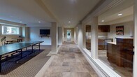 Located at 4215 S 30th St in Tacoma, Washington, this single-floor recreational facility is thoughtfully designed to accommodate a variety of leisure activities. The space is organized around a central tiled corridor that connects each distinct area, providing a clean and navigable layout throughout.

The billiards lounge serves as a focal point of the space, featuring a full-size pool table with a warm wood frame and red felt surface, illuminated by a decorative hanging fixture overhead. Bar-height stools line a wall-mounted shelf beside retro-style lounge signage and cue storage, lending the area a classic game-room character.

Adjacent to the billiards area is a comfortable media room, carpeted and fitted with a built-in wood entertainment unit housing a large flat-screen television alongside ample shelving and cabinetry. Leather recliner seating faces the screen, creating an ideal setting for relaxed viewing.

A dedicated ping-pong area provides additional recreational options, situated near the corridor and complemented by a wall-mounted television for added versatility. The kitchenette, visible through large glass partitions along the hallway, is equipped with cabinetry and countertops to support gatherings and events.

Throughout the space, recessed lighting and wall-mounted sconces maintain a warm, inviting ambiance, while neutral-toned carpet and stone-look tile flooring create a cohesive and polished aesthetic.