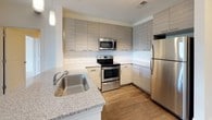 This single-floor, 2-bedroom, 2-bathroom residence combines modern design with functional living spaces throughout. The entryway opens to a well-lit foyer with dark hardwood-style flooring, multiple closets for ample storage, and clean white panel doors that carry consistently through the unit.

The living area features the same rich hardwood-style flooring and neutral walls, with a sliding glass door leading to a private balcony offering outdoor views of surrounding greenery. The kitchen is outfitted with contemporary flat-panel cabinetry in a light gray wood-grain finish, a white subway tile backsplash, granite countertops, stainless steel appliances including a refrigerator, range, and microwave, and a single-basin undermount sink.

Both bedrooms are carpeted in a light neutral tone and receive generous natural light through large windows. One bedroom includes a ceiling fan. The two bathrooms each reflect a clean, modern aesthetic. One features a granite-topped vanity, large-format white tile flooring, and a soaking tub, while the other offers a walk-in glass-enclosed shower with floor-to-ceiling white tile and brushed chrome fixtures. Together, the spaces present a cohesive, well-finished interior.