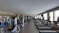 This modern fitness center is thoughtfully designed to accommodate a full range of workout routines. The space features smooth, neutral-toned walls, a clean dark floor surface, and a recessed lighting system that ensures the entire area is evenly and brightly illuminated. Large windows along one side of the facility allow natural light to fill the space, creating an open and energizing atmosphere. A row of True-brand treadmills lines the perimeter near the windows, complemented by additional cardio equipment to support endurance training. The strength training section is well-appointed with a multi-station cable and weight machine system, prominently featuring Hoist-branded equipment, along with a dedicated free weights area stocked with a full rack of dumbbells in varying sizes. Wall-mounted mirrors expand the visual depth of the space and allow users to monitor their form during workouts. A mounted flat-screen television is visible near the cardio section, adding to the amenity experience. The layout is spacious and thoughtfully organized, providing clear separation between cardio and strength zones while maintaining an uncluttered, professional environment suitable for users of all fitness levels.