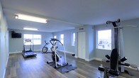 This single-floor fitness center offers a functional and well-organized space designed to support a range of workout routines. The room features light blue-grey walls paired with light hardwood-style flooring, creating a clean and energizing atmosphere. Multiple windows allow natural light to flow throughout the space, enhancing the open and airy feel of the interior. The fitness area is equipped with a variety of cardio machines, including a treadmill, stationary bike, and an elliptical trainer, each positioned to allow comfortable use and easy movement between equipment. Free weights and additional strength training equipment are also present, providing users with a comprehensive workout experience. Overhead fluorescent lighting ensures the space remains well-lit during all hours of use. A storage closet is conveniently located within the room, offering space for additional fitness accessories or supplies. The layout is thoughtfully arranged to maximize usable floor space while maintaining a safe and accessible environment. Overall, this fitness center presents a practical and inviting setting well-suited for daily exercise.