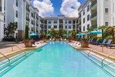 Located at 3450 Blair Cir NE in Brookhaven, Georgia, this beautifully designed outdoor amenity courtyard offers a resort-inspired retreat nestled within a multi-story residential community. The centerpiece of the space is a generously sized swimming pool with crystal-clear blue water, complemented by a waterfall feature and a surrounding brick-paved sun deck furnished with lounge chairs and teal umbrellas — ideal for relaxing outdoors.

Adjacent to the pool, a expansive flagstone patio provides a sophisticated gathering area anchored by a fully equipped outdoor kitchen. The kitchen features stainless steel grills, ample counter space with stone veneer finishes, and a pergola overhead — creating a welcoming space for outdoor cooking and dining. Nearby seating areas with bistro-style tables and cushioned chairs are shaded by umbrellas and framed by raised brick planters filled with tropical palms and manicured greenery.

Throughout the courtyard, tall palm trees and lush plantings lend a warm, resort-like ambiance, softening the architectural backdrop of the surrounding multi-story building. The combination of natural stone, brick detailing, and thoughtful landscaping creates a cohesive and inviting outdoor environment that encourages both social gatherings and peaceful relaxation.