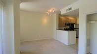 This single-floor residence offers an open and airy layout designed for comfortable everyday living. The living area is spacious and bright, featuring neutral-toned carpet flooring, crisp white walls, and elegant crown molding that adds a refined architectural detail throughout. A track lighting fixture overhead provides versatile illumination, enhancing the clean, well-maintained aesthetic of the space.

The kitchen flows seamlessly from the living area via a raised peninsula counter finished with dark granite countertops, creating a natural visual divider while maintaining an open feel. Warm-toned wooden cabinetry lines the kitchen walls, offering generous storage, while stainless steel appliances — including a microwave and refrigerator — add a modern, functional touch. The under-cabinet space and dedicated appliance area reflect a thoughtfully designed culinary workspace.

Visible ceiling vents suggest a central air system, ensuring year-round climate comfort. The combination of neutral finishes, crown molding, and the seamless transition between the living and kitchen spaces gives this residence a cohesive, move-in-ready ambiance that balances practicality with understated style.