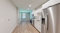 This thoughtfully designed single-floor unit offers a clean, modern aesthetic throughout its efficiently arranged layout. The open-concept main living area seamlessly integrates the kitchen and living space, all unified by light-toned hardwood-style flooring and neutral-toned walls that create a bright, airy atmosphere. Recessed lighting overhead enhances the sense of openness.

The kitchen is a standout feature, outfitted with flat-front white cabinetry extending to the ceiling for generous storage, a full suite of stainless steel appliances including a refrigerator with water dispenser, a gas range, built-in microwave, and dishwasher, as well as a single-basin undermount sink with a modern faucet set against a clean tile backsplash. A warm wood-toned entry door adds a natural contrast to the otherwise crisp white and gray palette.

The bedroom is carpeted for comfort and features a large black-framed window that admits natural light and provides views of a landscaped outdoor area. The bathroom is finished in an all-white scheme, featuring an undermount sink set in a white quartz vanity, a wall-mounted mirror, black matte hardware accents, and a glass-enclosed walk-in shower. The unit presents a cohesive, contemporary design that maximizes functionality within its compact footprint.