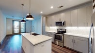 Located at 405 Crawford St in Fort Worth, TX, this modern single-floor unit offers a thoughtfully designed layout with high-quality finishes throughout. The open-concept kitchen serves as the heart of the space, showcasing gray flat-front cabinetry, white quartz countertops, and a full suite of stainless steel appliances including a range, microwave, and French door refrigerator. A central island with a deep undermount sink and industrial-style pendant lighting adds both function and style. A classic white subway tile backsplash and under-cabinet lighting complete the polished look, while a built-in wine rack provides an elegant storage accent. Rich, dark hardwood-style plank flooring flows throughout the main living areas, complemented by recessed lighting and a ceiling fan. The bedroom features plush carpet and a neutral color palette, creating a calm and comfortable retreat with natural light from a window. The bathroom offers a clean, contemporary design with dual-sink vanity topped in white quartz, sleek blue-gray cabinetry, a large framed mirror, and a glass-enclosed walk-in shower with tile surround. Large-format floor tiles tie the space together with a crisp, modern finish.