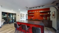 Located at 5415 Turtle Station in Westerville, Ohio, this single-floor community amenity space is designed with a vibrant, modern aesthetic that makes a strong visual impression throughout. The space is anchored by a standout wet bar area featuring a bold red subway tile backsplash and island facade, dark granite countertops, floating wood shelves stocked with glassware, and sleek track lighting overhead. Dark bar stools line the granite-topped island, creating an inviting social hub ideal for gatherings.

The open lounge area flows seamlessly from the bar and is appointed with wood-look plank flooring, a large wall-mounted flat-screen television, and a dramatic textured accent wall combining white geometric tile with bold red hexagonal elements. A full-length shuffleboard table serves as a central entertainment feature, complemented by plush sectional seating, a patterned area rug, a red ottoman, and an artisan chandelier. Curated abstract wall art adds a gallery-like quality throughout.

Natural light enters through large windows and a garage-style roll-up door that opens to an outdoor deck or patio, enhancing the connection between interior and exterior. Recessed lighting and clean white ceilings keep the space feeling bright and open. The overall ambiance is energetic yet comfortable, blending bold design choices with practical amenities for community use.