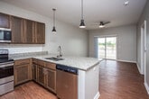 This well-appointed single-floor residence offers a thoughtfully designed open-concept layout that seamlessly connects the kitchen and living areas. The kitchen serves as the heart of the home, showcasing rich dark-toned cabinetry, granite countertops, and a classic white subway tile backsplash. A peninsula island with an undermount double sink and stylish pendant lighting provides additional prep space and a natural gathering point. Stainless steel appliances — including a refrigerator, range, microwave, and dishwasher — complete the kitchen's modern, functional aesthetic. Warm hardwood-style flooring flows consistently throughout the entire space, lending a cohesive and inviting feel. The living area is bright and open, illuminated by natural light from a sliding glass door that leads to an outdoor balcony area, offering views of the surrounding landscape. A ceiling fan adds both comfort and a finishing touch to the living space. The bedroom is a calm, neutral retreat featuring a window with a roller shade and its own ceiling fan. The bathroom is cleanly finished with granite-topped cabinetry, an undermount sink with brushed nickel fixtures, and hardwood-style flooring that maintains consistency with the rest of the home. Overall, the space presents a clean, contemporary design with quality finishes throughout.