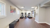 Located at 41 Highland Avenue in Randolph, Massachusetts, this fitness center offers a thoughtfully designed workout facility spread across two connected rooms on a single floor. The space is bright and welcoming, with flush-mounted ceiling light panels and multiple windows fitted with blinds that allow natural light to filter in throughout the day.

The primary cardio room features carpeted flooring and houses a collection of treadmills and an elliptical machine, providing ample space for cardiovascular training. A sleek bench lines one wall alongside decorative artwork, contributing to the clean, modern aesthetic maintained throughout. An analog wall clock and an emergency exit sign underscore the practical, community-focused design.

The second room transitions to light hardwood-style flooring and is dedicated to strength training. It is equipped with a full dumbbell rack offering a range of weights, a multi-function cable resistance machine, and a spin bike, making it suitable for a comprehensive strength and cross-training workout. Motivational wall art and additional windows add energy and airiness to the space.

Together, the two rooms form a compact yet versatile fitness facility. The clean white walls, consistent trim work, and organized equipment layout create a polished and functional environment well suited for everyday exercise.