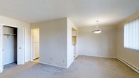 This single-floor residence offers a practical and well-organized layout designed for comfortable living. The main living area is carpeted in a neutral tone, providing a warm and understated foundation that pairs naturally with the soft beige walls throughout. A pendant light fixture hangs overhead, adding a simple decorative touch while providing focused illumination to the space.

The kitchen is semi-open, separated from the living area by a partial wall that maintains a sense of openness while defining each zone. White cabinetry lines the kitchen walls, complemented by white appliances including a refrigerator and stove, contributing to a clean and cohesive aesthetic. The kitchen features hard flooring, offering a practical contrast to the carpeted living area.

The textured ceiling runs consistently throughout, adding subtle visual interest overhead. The neutral color palette of whites and beiges across walls, cabinetry, and flooring creates a bright, airy ambiance that makes the space feel open and easy to personalize. Overall, the residence presents a straightforward and functional environment suited to a range of lifestyles.