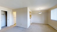 This single-floor residence offers a practical and well-organized layout designed for comfortable living. The main living area is carpeted in a neutral tone, providing a warm and understated foundation that pairs naturally with the soft beige walls throughout. A pendant light fixture hangs overhead, adding a simple decorative touch while providing focused illumination to the space.

The kitchen is semi-open, separated from the living area by a partial wall that maintains a sense of openness while defining each zone. White cabinetry lines the kitchen walls, complemented by white appliances including a refrigerator and stove, contributing to a clean and cohesive aesthetic. The kitchen features hard flooring, offering a practical contrast to the carpeted living area.

The textured ceiling runs consistently throughout, adding subtle visual interest overhead. The neutral color palette of whites and beiges across walls, cabinetry, and flooring creates a bright, airy ambiance that makes the space feel open and easy to personalize. Overall, the residence presents a straightforward and functional environment suited to a range of lifestyles.