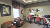 This single-floor space combines functional design with a warm, inviting ambiance. The business center area is thoughtfully equipped with a granite-topped workstation accommodating two computer workstations, complete with ergonomic rolling chairs upholstered in a muted rose tone. Framed artwork adorns the walls above the desk, adding a polished, professional touch to the workspace.

Adjacent to the workstation, a comfortable lounge seating area features upholstered armchairs with striped accent cushions in complementary earth tones, ideal for casual meetings or quiet relaxation. A wall-mounted roman shade in a woven striped pattern filters natural light through the window, creating a soft, diffused glow across the neutral-carpeted floor.

The tray ceiling with recessed lighting adds architectural depth to the room, while warm beige and gray wall tones contribute to an overall calming atmosphere. Decorative accents, including a wall clock, a tall floral arrangement, and a magazine rack, enhance the space's approachable character. The cohesive design balances practicality with comfort, making this space well-suited for residents or visitors seeking a productive yet relaxed environment.