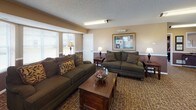 This single-floor communal space is thoughtfully designed to foster comfort and social interaction across several well-defined areas. The main lounge is generously proportioned, featuring plush dark-green upholstered sofas accented with decorative pillows, a rich wood coffee table, and warm ambient lighting from table lamps positioned throughout. A two-tone wall treatment — tan on the upper half and deep brown on the lower — lends a grounded, traditional aesthetic that runs consistently throughout the space. Natural light filters in through multiple windows with blinds, offering views to the outside while maintaining a cozy interior atmosphere. A textured, patterned carpet covers the floors in the main living and dining areas, adding warmth underfoot. The media and seating area is anchored by a flat-screen television and additional sectional seating, making it well-suited for group gatherings. Adjacent to the lounge, a dedicated dining area accommodates a full wooden dining table with chairs, positioned near a breakfast bar with counter seating. The attached kitchenette is equipped with white cabinetry, a full-size refrigerator, a sink, and countertop workspace, all set atop durable hardwood-style flooring. Overall, the space presents a welcoming, functional environment ideal for community use.
