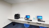 This single-floor amenity space is divided into two distinct functional areas, each thoughtfully designed to meet the needs of its users. The business center features continuous wall-mounted white countertop workstations that run along multiple walls, providing ample desk space for individual use. Multiple desktop computers with monitors and keyboards are stationed throughout, along with a multifunction printer for scanning, copying, and printing needs. Ergonomic blue rolling stools on a patterned blue-and-grey carpet add a clean, modern aesthetic to the bright, white-walled room with a drop-panel ceiling. The fitness center is equally well-appointed, featuring a durable rubberized floor and a comprehensive selection of cardio and strength equipment. Treadmills, ellipticals, a stationary bike, a multi-station cable machine, adjustable benches, and a full dumbbell rack are all available. Wall-mounted televisions provide entertainment during workouts, while large windows look out toward a pool area, filling the space with natural light and outdoor views. A warm wood-accent wall adds a design focal point, complementing the industrial-style exposed ductwork and overhead fluorescent lighting throughout.