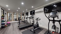 This single-floor fitness studio is thoughtfully designed to accommodate both active workouts and comfortable relaxation. The layout is organized into two distinct zones: a fully equipped gym area and a welcoming lounge space.

The gym area is finished with light wood-look luxury vinyl flooring and features white shiplap accent walls that give the space a clean, modern farmhouse aesthetic. Recessed lighting throughout ensures bright, even illumination across the workout area. Equipment includes a treadmill, spin bike, elliptical, adjustable weight bench, and an Olympic barbell rack with weights. Multiple wall-mounted flat-screen televisions and a wall-mounted mini-split air conditioning unit enhance the workout experience. A series of large, wood-framed mirrors line one wall, visually expanding the space and serving as functional training aids. A medicine ball rack adds additional strength training options.

The lounge area transitions seamlessly from the gym, featuring light-toned flooring and neutral gray walls. Two chartreuse mid-century modern accent chairs with channel-tufted upholstery flank a sculptural live-edge wooden side table topped with a potted plant, creating a warm and inviting setting for rest and recovery.
