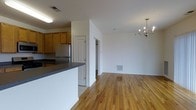 This single-floor space offers an open-concept design that seamlessly connects the kitchen and living area. The kitchen features warm honey-toned wood cabinetry, a stainless steel refrigerator, and a peninsula with a dark countertop that provides additional workspace and serves as a natural divider between the cooking and living zones. Tile flooring anchors the kitchen area, while rich hardwood floors extend throughout the main living space, adding warmth and continuity to the layout. The living area is generously proportioned, with neutral white walls that create a clean, versatile backdrop. A decorative chandelier serves as a focal point overhead, lending a touch of elegance to the open room. Visible ductwork registers suggest a central HVAC system throughout. A doorway visible from the main area hints at additional rooms or storage beyond, contributing to the functional flow of the floor plan. The overall ambiance is bright and understated, offering a practical yet inviting environment well-suited for everyday living.