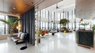 The Sky Lounge in Coquitlam, BC is a thoughtfully designed rooftop amenity space that blends contemporary architecture with eclectic, vibrant interiors across a generously sized single floor. The main lounge is defined by soaring double-height ceilings and an expansive curtain wall of floor-to-ceiling windows that floods the space with natural light and frames sweeping panoramic views of the surrounding cityscape and mountains. Gleaming terrazzo floors, ceiling fans, and a dramatic circular sectional sofa centered around a large indoor tree create a striking focal point in this open-plan gathering area. Ornate starburst partition screens divide the space with visual interest, while designer seating—including iconic swan chairs and tufted leather daybeds—adds a mid-century modern character. A dedicated bar area features a beautifully crafted counter clad in teal geometric tiles beneath warm wood-paneled ceilings and brass pendant lighting. An adjoining lounge room offers a more intimate, bohemian atmosphere complete with tropical leaf murals, rattan furniture, herringbone wood floors, and a karaoke setup with neon signage. A glass-enclosed outdoor terrace provides additional al fresco seating with open-air views, completing this versatile and stylish sky-high retreat.