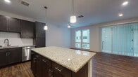 This single-floor residence offers a thoughtfully designed open-concept layout that seamlessly connects the kitchen, dining, and living areas. The kitchen is a standout feature, showcasing dark espresso cabinetry, a white subway tile backsplash, stainless steel appliances including a refrigerator, range, microwave, and dishwasher, and a large central island topped with light granite countertops. Pendant lighting above the island adds a modern touch, while recessed lighting illuminates the space throughout. Rich hardwood-style flooring extends across the main living areas, complementing the contemporary color palette of dark cabinetry against crisp white walls.

The living area benefits from abundant natural light through large windows dressed with vertical blinds, and a ceiling fan contributes to the airy, comfortable atmosphere. The bedrooms are carpeted for warmth and comfort, each featuring windows that invite natural light. One room includes built-in shelving, offering convenient storage solutions alongside hardwood-style flooring.

The bathroom is finished to a high standard, with a granite-topped dark vanity, a framed mirror, tile flooring, and a full tub-shower combination with white subway tile surround. Overall, the unit presents a cohesive, modern aesthetic with quality finishes throughout.