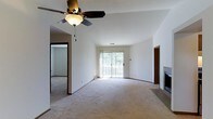 This single-floor space centers around a generously sized living area that is both open and inviting. The room is finished with wall-to-wall neutral carpeting, which adds warmth and comfort underfoot. A ceiling fan with a light fixture is mounted overhead, providing both air circulation and ambient lighting, while contributing to the room's clean, functional aesthetic.

A sliding glass door dominates one wall, flooding the space with natural light and offering a view of an outdoor deck surrounded by mature trees. This seamless connection between the interior and exterior creates an airy, bright atmosphere throughout the living area. A fireplace is positioned along an adjacent wall, serving as a notable architectural feature that adds character and a potential focal point for the room.

The layout includes a connected secondary room or hallway visible through a doorway, suggesting a well-organized floor plan with clear room separation. The white walls throughout maintain a neutral, versatile backdrop, making the space adaptable to a variety of interior design preferences. Overall, the space presents a comfortable, functional layout with thoughtful natural light integration.