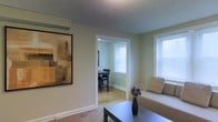 This single-floor residence offers a comfortable and well-appointed layout designed for everyday living. The living area is finished with soft carpeting underfoot and features a textured ceiling that adds subtle depth to the room. A large framed abstract artwork adorns one wall, lending an artistic touch to the space, while a sofa with accent pillows and a sleek coffee table complete the furnishings. Natural light filters in generously through a window fitted with horizontal blinds, creating a bright and welcoming atmosphere. A wide, open doorway connects the living area to the dining room, which features warm hardwood flooring that contrasts nicely with the carpeted living space. The dining room accommodates a dark wood table and chairs, and benefits from its own window that brings in additional natural light. The walls throughout are finished in a soft, neutral green tone that contributes to a calm and cohesive ambiance. Baseboard heating elements are visible along the walls, indicating practical year-round comfort. The open flow between the living and dining areas makes the overall layout feel connected and spacious, ideal for both relaxed everyday use and casual entertaining.