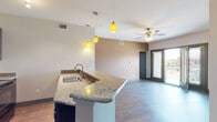 This single-floor residence offers a well-designed open-concept layout that seamlessly connects the kitchen, dining, and living areas. The kitchen is outfitted with dark espresso cabinetry, granite countertops, and a full suite of black appliances including a refrigerator, range, and built-in microwave. A peninsula with an integrated sink and striking yellow pendant lights serves as a natural divider between the kitchen and the main living space, which features wood-style flooring and a ceiling fan. Sliding glass doors open to a private balcony, allowing natural light to fill the living area and providing outdoor views. The bedrooms are carpeted and well-lit, each equipped with ceiling fans and horizontal blinds covering windows that bring in additional natural light. The bathroom includes a full tub-shower combo with large-format tile surround, wood-look flooring, granite-topped dark cabinetry, and modern brushed nickel fixtures. Throughout the residence, a consistent design palette of neutral tones, dark trim, and contemporary finishes creates a cohesive and polished aesthetic. The thoughtful floor plan maximizes functionality while maintaining an open, airy feel across all living spaces.