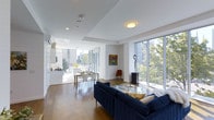 This single-floor space showcases a modern open-plan layout that seamlessly connects the living, dining, and kitchen areas into one cohesive, light-filled environment. Large floor-to-ceiling windows line the exterior walls, flooding the interior with abundant natural light and framing lush treetop and urban skyline views. The crisp white walls and tray ceilings enhance the sense of openness and volume throughout.

The living area is anchored by a deep navy blue sofa set around a glass coffee table, complemented by curated artwork and a sleek sculptural accent piece. Warm hardwood flooring runs throughout, adding a rich contrast to the bright, contemporary palette. A compact dining area with a clean white table and modern chairs sits adjacent to the kitchen, which features white cabinetry, a gas range, and a minimalist design that maintains the overall aesthetic continuity.

Natural light, an open layout, and large windows create an airy, welcoming ambiance, making the space feel both functional and refined. The overall design balances modern minimalism with warm, livable details.