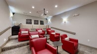 Located at 1450 Bluewater Way in Charleston, SC, this dedicated screening room delivers a refined, intimate theater experience within a compact yet thoughtfully designed space. The room is anchored by a ceiling-mounted projector paired with a large pull-down projection screen framed by elegant floor-length curtains, creating a true cinematic focal point. Seating is a standout feature, combining plush red leather club chairs arranged around small circular side tables with a tufted cream sofa along one wall, offering a variety of comfortable viewing options. Decorative film reel artwork in wooden frames lends a classic Hollywood charm to the neutral gray walls, which are warmly illuminated by stylish wall sconces. The patterned carpet — a light, speckled design — adds texture and visual interest underfoot. Recessed ceiling lighting supplements the ambient sconces, ensuring the room is well-lit when not in screening mode. A handrail along one side wall suggests gentle tiered access, maximizing sightlines for all seats. The overall ambiance is polished and inviting, blending the excitement of a movie theater with the comfort of a premium lounge, making it an ideal venue for private screenings and social gatherings.