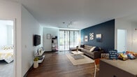 This single-floor residence presents a thoughtfully designed layout with a contemporary aesthetic throughout. The living room serves as the focal point of the space, distinguished by a striking deep navy blue accent wall adorned with a curated trio of framed artwork. A large sectional sofa anchors the room, complemented by a sleek white coffee table and a patterned area rug. Large sliding glass doors flood the space with natural light and provide access to an outdoor area, offering views of greenery beyond. Track lighting on the ceiling adds a modern, functional touch.

The kitchen is outfitted with stainless steel appliances, including a full-size refrigerator, range, microwave, and dishwasher. White upper cabinetry contrasts with dark lower cabinets, and a kitchen island provides additional prep and dining space. Wood-tone flooring runs throughout the kitchen and living areas, adding warmth to the modern palette.

The bedroom is a calm, neutral retreat with soft grey walls, a bright window with blinds, and space for a desk and study area. Two bathrooms serve the unit — one featuring a glass-enclosed shower and a dark-cabinetry vanity, and the other a streamlined vanity with clean, minimal finishes throughout.