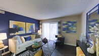This single-floor living space showcases a thoughtfully designed interior that balances bold color choices with a comfortable, contemporary aesthetic. The living area serves as the focal point of the space, anchored by a striking navy blue accent wall that provides a dramatic backdrop for the room's decor. Two large, colorful abstract paintings hang above a neutral-toned sofa dressed with an array of decorative pillows in coordinating shades of blue, yellow, and grey.

Dark hardwood-style flooring runs throughout the space, offering a rich contrast to the lighter furnishings. A geometric-patterned area rug in navy and white defines the seating area, which is further complemented by a sleek dark coffee table and a pair of rounded grey leather accent chairs with nailhead trim detailing.

A full-length sliding glass door dominates one wall, allowing generous natural light to flow into the space and providing a direct view of the outdoors. Striped curtain panels frame the door, adding visual height to the room. The textured ceiling completes the space, contributing to its layered, well-appointed character. Overall, the space presents a cohesive and stylish living environment with a vibrant, modern personality.