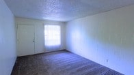 This single-floor space presents a straightforward and functional layout suited for everyday living. The main room is finished with wall-to-wall carpeting that adds warmth underfoot, while a textured popcorn ceiling runs overhead, giving the space a classic residential feel. One wall features wood panel detailing, providing a subtle contrast to the otherwise neutral surroundings. Natural light enters through a window dressed with vertical blinds, which can be adjusted to control light and privacy throughout the day. A doorway is visible, offering easy access to the adjoining areas of the space. The overall ambiance is clean and understated, providing a blank canvas that can be personalized to suit a variety of needs and preferences. The open, uncluttered nature of the room allows for flexible furniture arrangements, making it adaptable for use as a living area or multi-purpose room. The combination of carpeted flooring, paneled walls, and diffused natural light creates a calm, comfortable atmosphere throughout this single-level space.