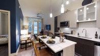 This single-floor space showcases an open-concept layout that seamlessly integrates the kitchen, dining, and living areas into one cohesive, functional environment. The kitchen is appointed with dark cabinetry contrasted by white upper cabinets and light stone countertops, complemented by a subway tile backsplash and stainless steel appliances, including a built-in microwave. Pendant lighting hangs above the kitchen island, which doubles as an extended dining surface, providing both prep space and casual seating. The dining area features a marble-topped table with leather chairs, adding a refined yet approachable aesthetic. Hardwood-style flooring runs throughout, lending warmth and visual continuity across the space. The living area is positioned near large French-style windows that invite abundant natural light and offer views of the outdoor surroundings. A ceiling fan is installed overhead, enhancing comfort. The overall ambiance balances modern design with warm, inviting tones, creating a space that is both practical and aesthetically well-considered.
