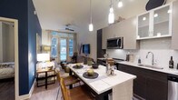 This single-floor space showcases an open-concept layout that seamlessly integrates the kitchen, dining, and living areas into one cohesive, functional environment. The kitchen is appointed with dark cabinetry contrasted by white upper cabinets and light stone countertops, complemented by a subway tile backsplash and stainless steel appliances, including a built-in microwave. Pendant lighting hangs above the kitchen island, which doubles as an extended dining surface, providing both prep space and casual seating. The dining area features a marble-topped table with leather chairs, adding a refined yet approachable aesthetic. Hardwood-style flooring runs throughout, lending warmth and visual continuity across the space. The living area is positioned near large French-style windows that invite abundant natural light and offer views of the outdoor surroundings. A ceiling fan is installed overhead, enhancing comfort. The overall ambiance balances modern design with warm, inviting tones, creating a space that is both practical and aesthetically well-considered.
