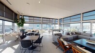 This contemporary amenity lounge is defined by its expansive floor-to-ceiling windows that wrap around the perimeter, flooding the interior with natural light and offering sweeping panoramic views of the surrounding cityscape. The open-plan layout is generously proportioned, with large-format tile flooring and recessed lighting that contribute to a clean, sophisticated aesthetic throughout.

The main lounge area is thoughtfully arranged to accommodate a variety of uses. A plush sectional sofa and mid-century modern accent chairs are anchored by a dark-veined area rug, creating an inviting social hub. Nearby, multiple dining tables with woven chairs offer flexible seating for casual gatherings or work sessions. A billiards table serves as a central recreational feature, while a wall-mounted television adds to the entertainment options. Lush potted plants punctuate the space, softening the modern architecture with organic warmth.

A dedicated kitchenette area features light wood cabinetry with a white countertop, an integrated sink, a stainless steel refrigerator, and floating wood shelves styled with decorative accessories. Restrooms are conveniently accessible from this area. Roller shades on all windows allow for adjustable light control while preserving the connection to the stunning outdoor views and adjacent terrace, making this a highly functional and visually striking communal space.
