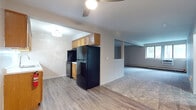 This upgraded two-bedroom, one-bathroom unit is laid out across a single floor, offering a well-organized and functional living space. The galley-style kitchen is a standout feature, equipped with warm honey-toned wood cabinetry, light countertops, a stainless steel sink with a modern faucet, and black appliances including a refrigerator, range, and dishwasher. Durable wood-look vinyl plank flooring runs throughout the kitchen, while a decorative chandelier adds a refined touch overhead. The spacious living area is carpeted and bathed in natural light through large windows that frame outdoor greenery. A ceiling fan adds comfort, and baseboard heating is present throughout. A half-wall partition separates the kitchen from the living space, creating an open yet defined layout. Both bedrooms are carpeted and neutrally painted, offering quiet and versatile spaces with ample room for furnishings. Built-in shelving is visible near one bedroom entry, providing convenient storage. The bathroom has been thoughtfully updated with a grey shaker-style vanity, white countertop, wood-look vinyl flooring, chrome fixtures, and a tiled tub-shower combination. The overall aesthetic is clean, neutral, and move-in ready.