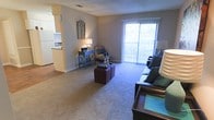 This single-floor residence offers a comfortable and functional layout designed for everyday living. The living area serves as the heart of the space, featuring plush wall-to-wall carpeting that adds warmth underfoot. A large sliding glass door floods the room with natural light and opens to a private balcony, providing a pleasant outdoor connection and views of the surrounding area. The open layout creates a seamless flow between the living and dining areas, making the space feel airy and inviting. Neutral-toned walls complement the carpeting and provide a versatile backdrop for a variety of décor styles. A decorative accent wall area adds visual interest and a touch of character to the living room. Adjacent to the living space, the kitchen is partially open to the main area, allowing for easy interaction between rooms. The kitchen features light cabinetry and includes essential appliances, offering a practical and efficient cooking environment. Recessed ceiling vents suggest a central HVAC system for year-round climate comfort. Overall, the space presents a clean, well-proportioned layout with a cozy ambiance, ample natural light, and functional design elements that make it a comfortable and livable environment.