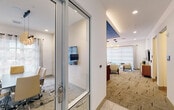 This thoughtfully designed single-floor amenity space combines professional functionality with refined aesthetics, offering multiple distinct zones suited for both collaborative work and relaxed gatherings. The layout is anchored by a glass-walled conference room accessible through sleek aluminum-framed double doors, providing transparency while maintaining acoustic separation. Inside, a dark wood conference table is surrounded by cream and gold upholstered chairs, illuminated by a sculptural pendant light fixture and a large wall-mounted display screen. Natural light filters in through sheer-curtained windows, enhancing the room's professional yet welcoming atmosphere.

Adjacent to the conference room, a main lounge area features warm, neutral tones with patterned carpet flooring and a wall-mounted television atop a light wood credenza. Two private pod rooms, distinguished by bold geometric-patterned walls, offer intimate spaces for focused use. A separate seating nook with floor-to-ceiling windows wrapped in roller shades invites abundant natural light, furnished with cream leather armchairs, wood-accented side tables, and a textured ottoman beneath striking circular gold pendant lights. The overall design balances modern sophistication with functional versatility.
