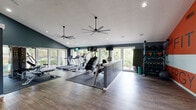 Located at 9821 Summerwood Cir in Dallas, TX, this fitness center offers a well-appointed, single-floor workout facility designed to support a comprehensive exercise experience. The space features vaulted ceilings that lend an open, airy feel, complemented by large ceiling fans that ensure comfortable airflow throughout. Recessed lighting, combined with an abundance of natural light streaming through floor-to-ceiling windows, creates a bright and energizing atmosphere with pleasant views of the surrounding greenery.

The cardio zone is outfitted with multiple treadmills, an elliptical, a stair climber, and additional cardio machines, all positioned to take advantage of the outdoor views. A dedicated strength training area features a multi-station cable machine, a full rack of dumbbells, and adjustable benches, providing versatile options for resistance workouts.

The interior is finished with durable hardwood-style flooring and rubber mats in workout zones for safety and comfort. Deep teal-blue walls anchor the color palette, while a bold accent wall in orange and gray displays motivational words — "Fit," "Focus," "Exceed," and "Strength" — adding an energetic visual statement. Glass-paneled entry doors and a mirrored section enhance the sense of space. The overall design balances function and aesthetics, creating a motivating environment for fitness.