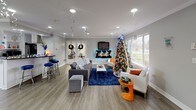This single-floor clubhouse is a thoughtfully designed community space that seamlessly blends functionality with a warm, inviting atmosphere. The main lounge area serves as the heart of the clubhouse, featuring wood-look flooring, crown molding, and recessed lighting throughout. A wall-mounted flat-screen TV is positioned above a fireplace mantle, flanked by comfortable seating arrangements including a plush gray sofa, accent chairs, and a bold blue area rug that anchors the space. Large windows flood the room with natural light while offering pleasant views of the surrounding greenery outdoors. A bar-height seating area with vibrant blue stools provides an additional social gathering spot along one wall. Adjacent to the lounge, a fully equipped kitchen offers ample cabinetry, granite countertops, stainless steel appliances, and a central island. A bistro-style dining table with bar-height chairs sits near sliding glass doors that open to an outdoor patio, enhancing the connection between interior and exterior spaces. A welcoming reception nook features a small counter with a coffee station and a chalkboard, directing residents toward a private office area equipped for administrative needs. The cohesive gray-and-white color palette, crown molding details, and consistent flooring throughout create a polished and unified aesthetic across all areas of this versatile clubhouse.